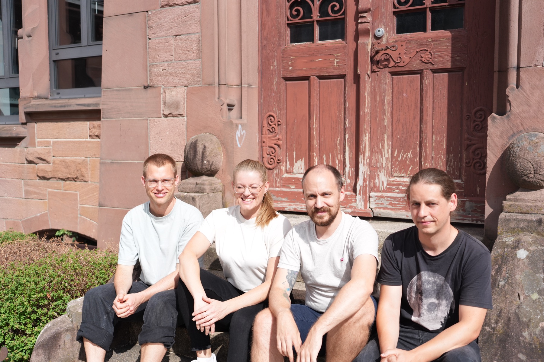 Lars Arendt, Sarah Huber, Marvin Bernemann und Matthias Windeisen sitzen nebeneinander auf einer Treppenstufe vor einem alten Sandsteinportal.